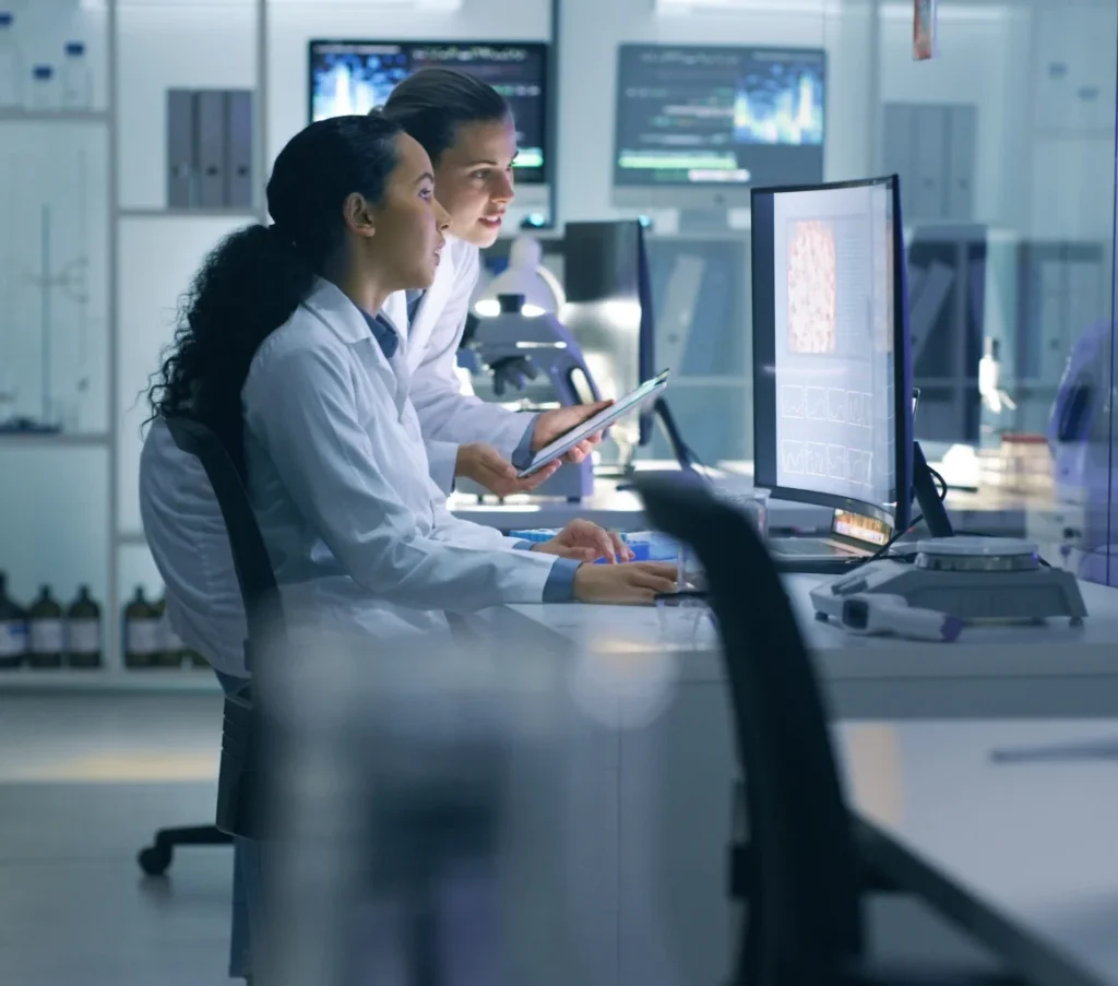 Two female scientists in lab coats analyzing data on a computer and tablet in a modern laboratory.