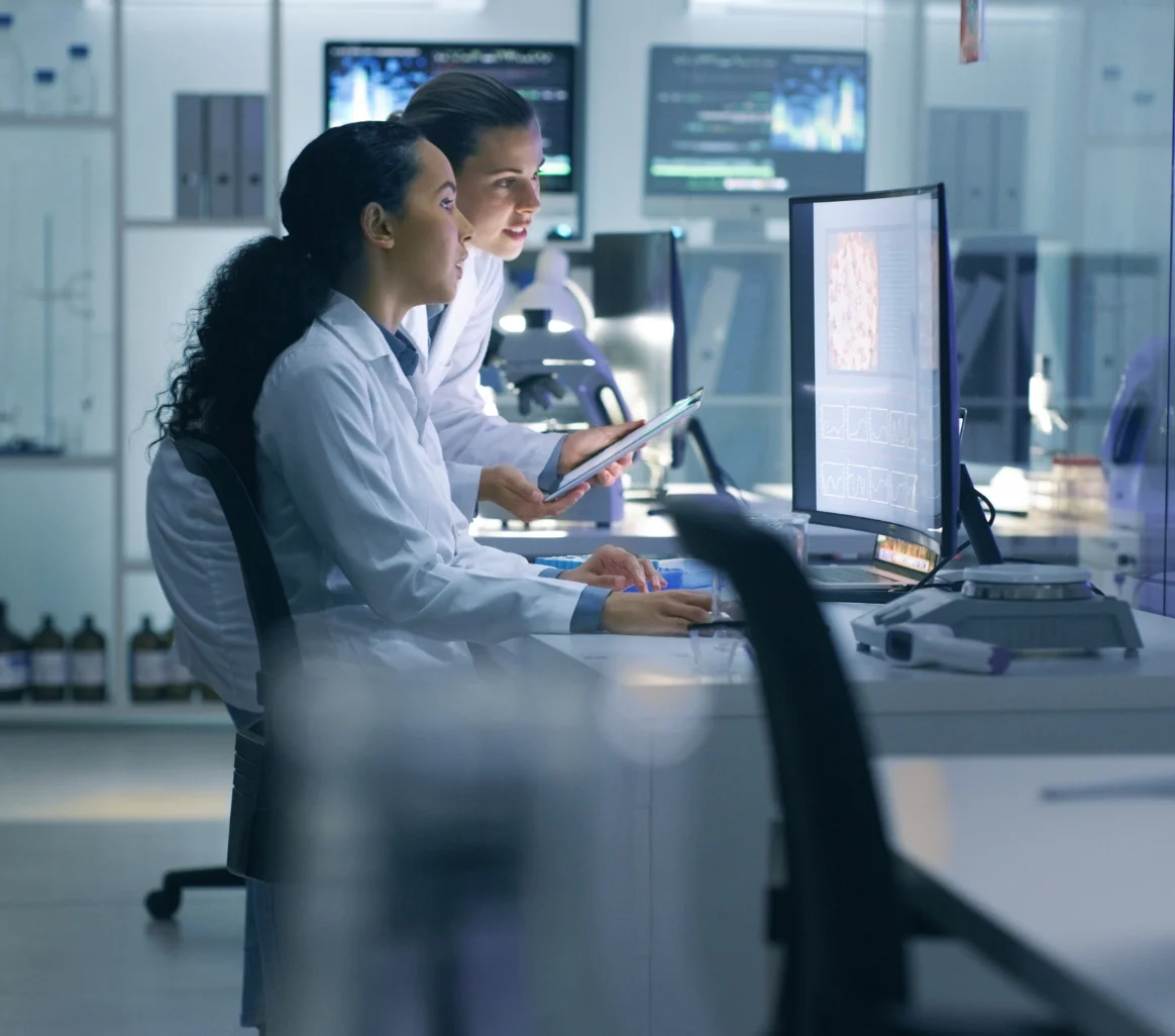 Two female scientists in lab coats analyzing data on a computer and tablet in a modern laboratory.
